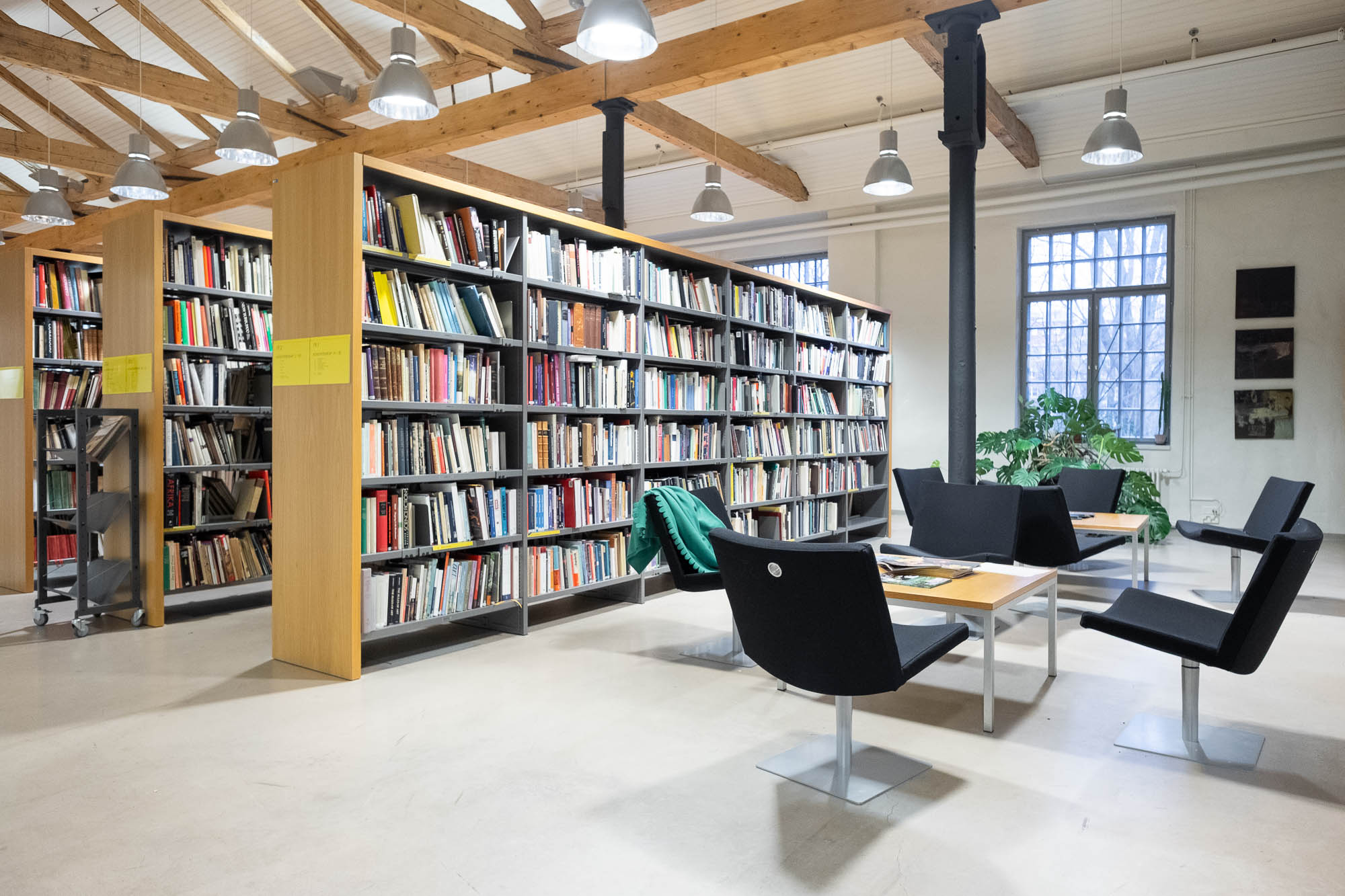 A well-lit library interior with tall bookshelves filled with books, several black chairs around tables, large windows letting in daylight, and exposed wooden ceiling beams.