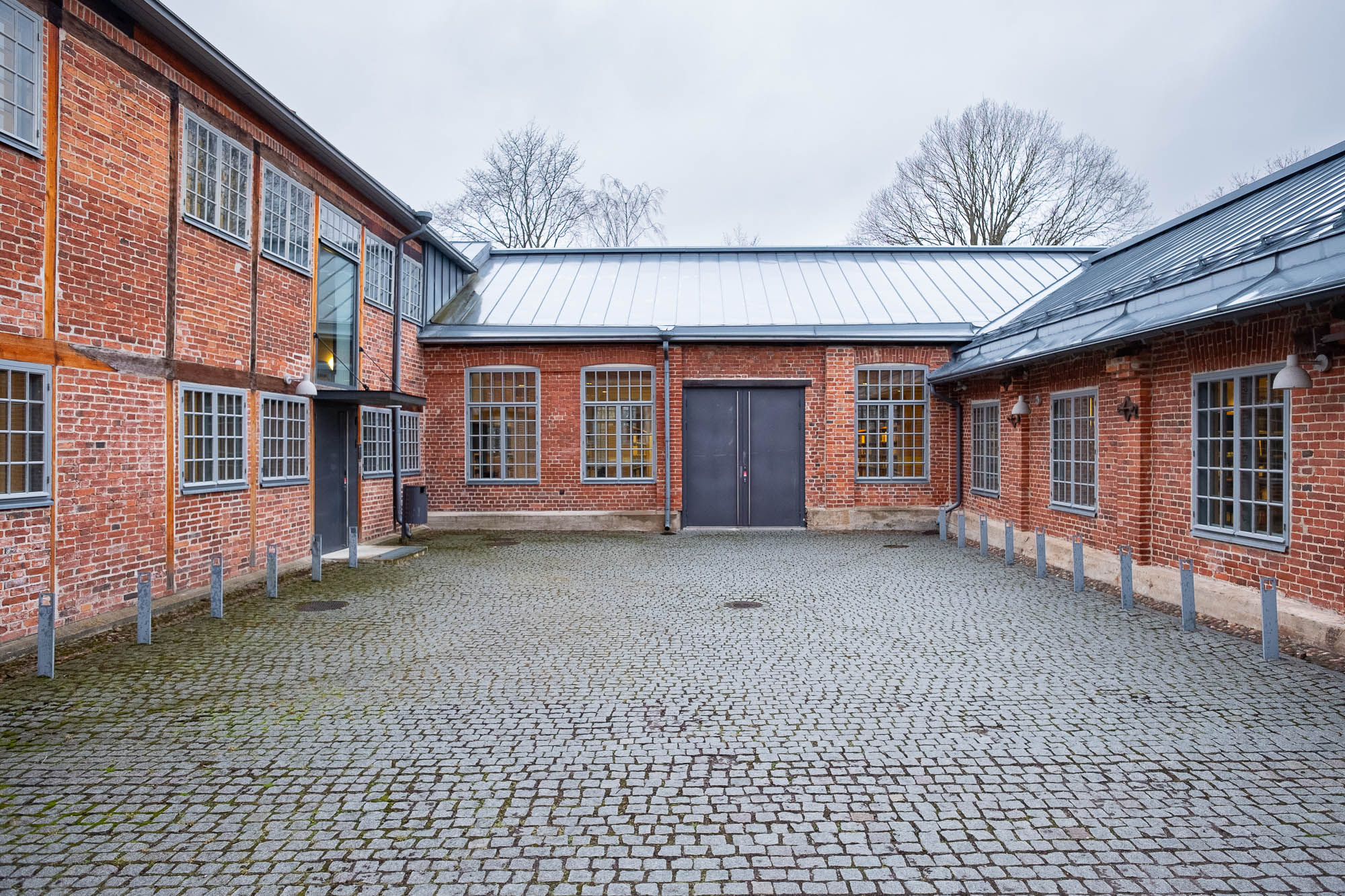 A paved courtyard surrounded by red brick buildings with large grid windows and metal roofs on a cloudy day. There are some trees visible in the background behind the buildings.