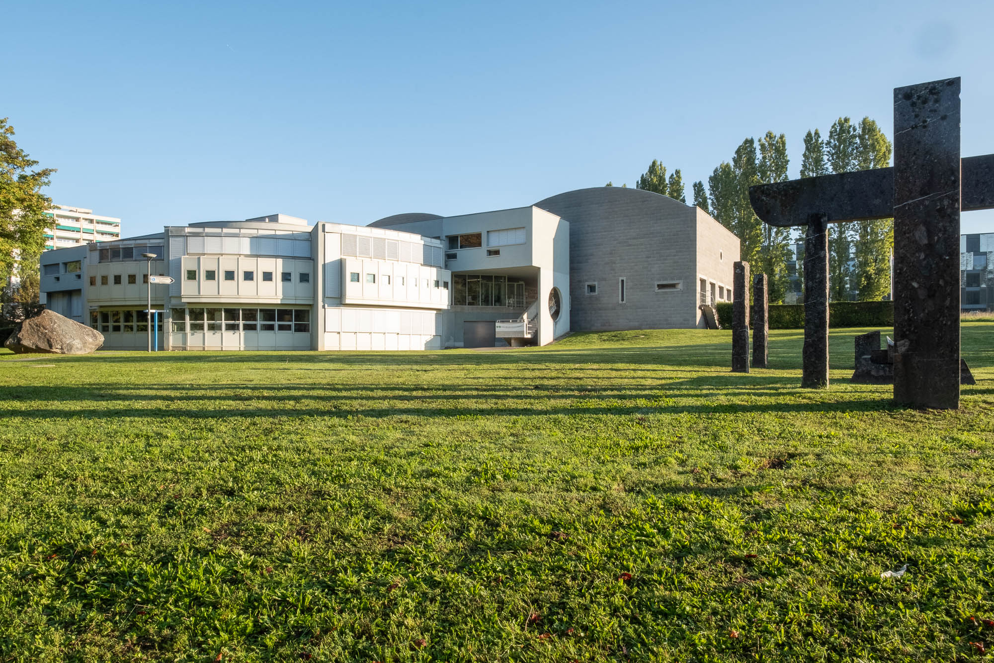 Exterior of a postmodern building, comprised of curved sections fanning out towards a grass meadow. To the right is a sculpture made out of geometric stone pieces.