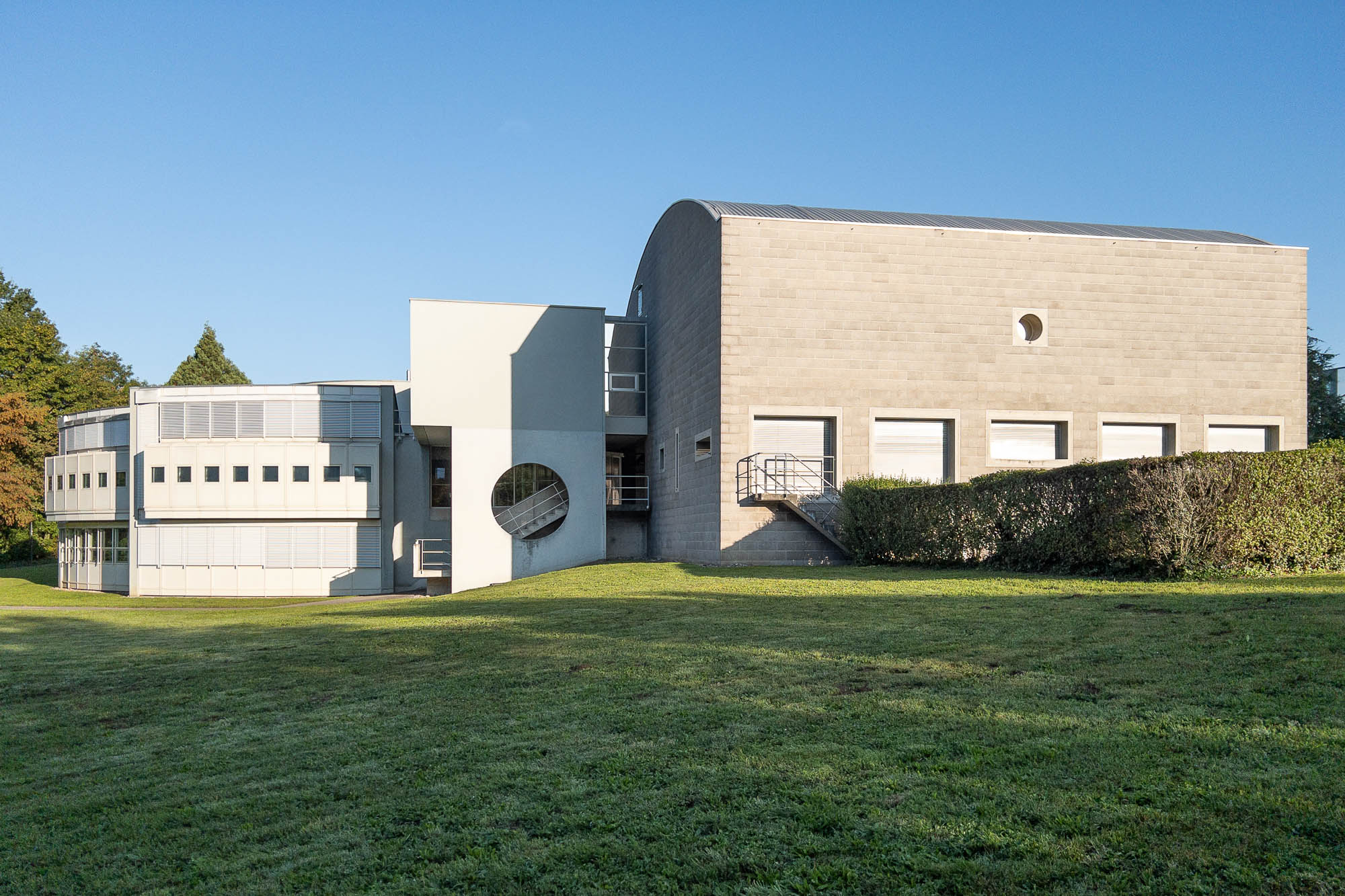 A postmodern building with a minimalist design, featuring a rectangular section with small windows and a curved roof, connected to a lighter-colored wing with geometric shapes, set on a grassy lawn under a clear blue sky.