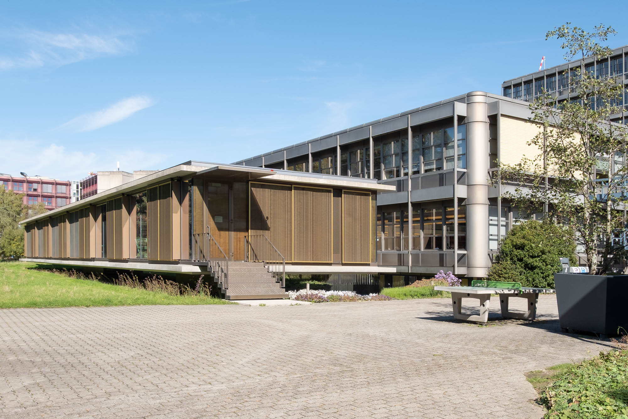 A modern, two-story building with large windows and brown panels, surrounded by grass and trees, under a clear blue sky. Concrete steps lead to the entrance, and a paved area is in the foreground.