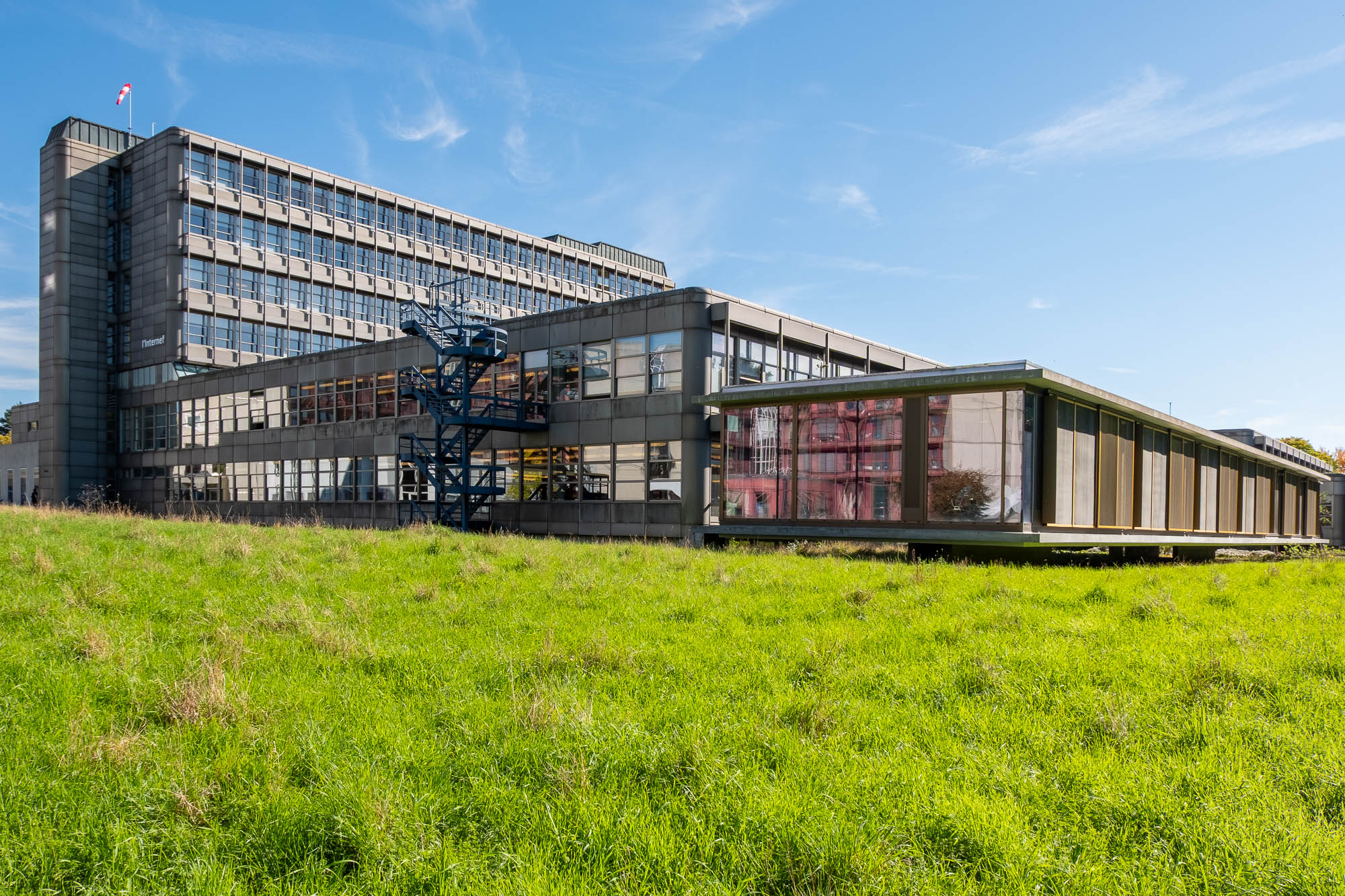 A large, modern building with many windows and an external metal staircase sits beside a smaller glass-walled structure, both surrounded by a grassy field under a clear blue sky.