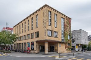 A three-story yellow library building on a street corner with large vertical windows, bicycles parked outside, ivy growing on one side, and people walking nearby. Signs on the facade read "Stadtbibliothek Bibliothèque de la Ville Biel / Bienne".