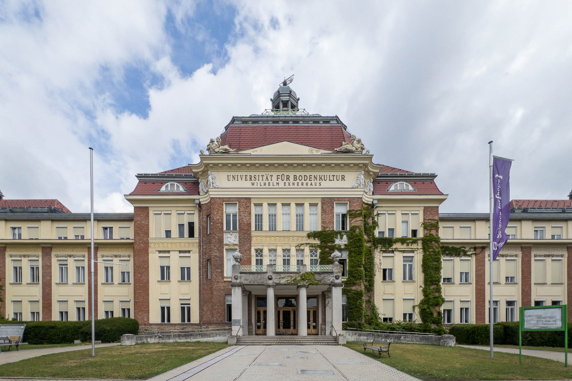 Front view of a large, historic university building with a red roof, yellow and brick facade, and central entrance. Universität für Bodenkultur is written above the entrance. Two flagpoles stand in front.