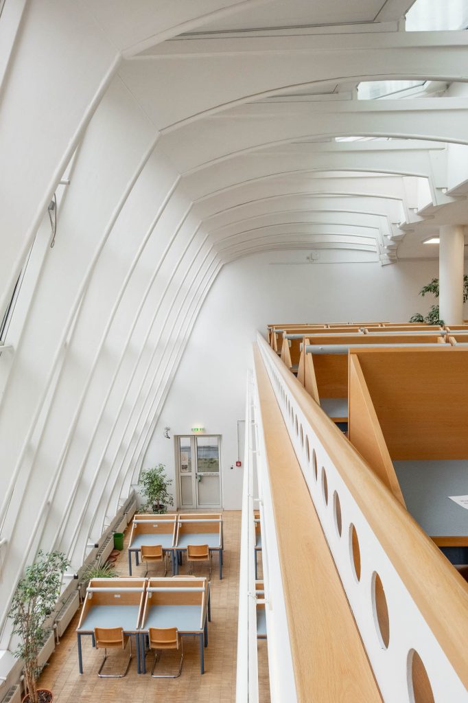 A modern library interior with high arched ceilings, white walls, rows of wooden desks and chairs, potted plants, and a mezzanine level with a wooden railing featuring circular cutouts.