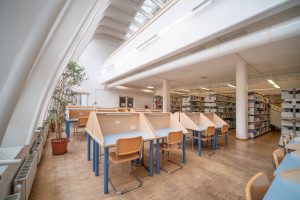 A modern library interior with wooden study desks separated by partitions, wooden chairs, bookshelves, a tall potted plant, large windows, and high white ceilings with skylights.