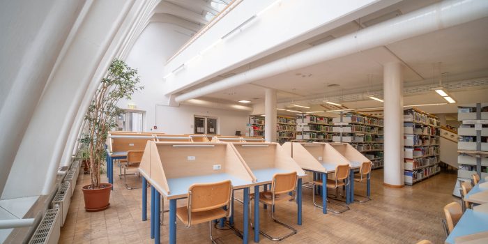 A modern library interior with wooden study desks separated by partitions, wooden chairs, bookshelves, a tall potted plant, large windows, and high white ceilings with skylights.