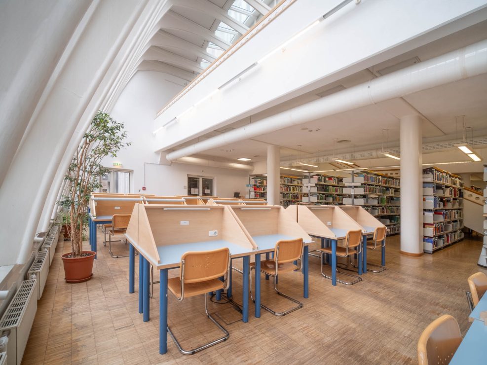 A modern library interior with wooden study desks separated by partitions, wooden chairs, bookshelves, a tall potted plant, large windows, and high white ceilings with skylights.