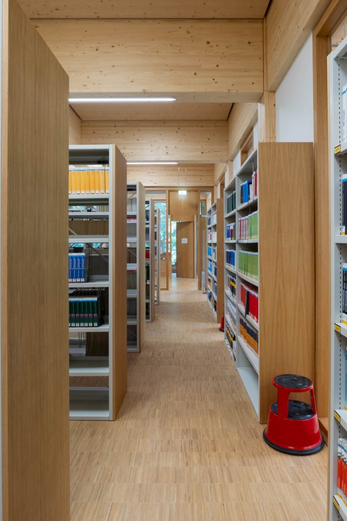 A library aisle with tall wooden shelves filled with books, a wooden floor, and a red step stool positioned near the right shelves.
