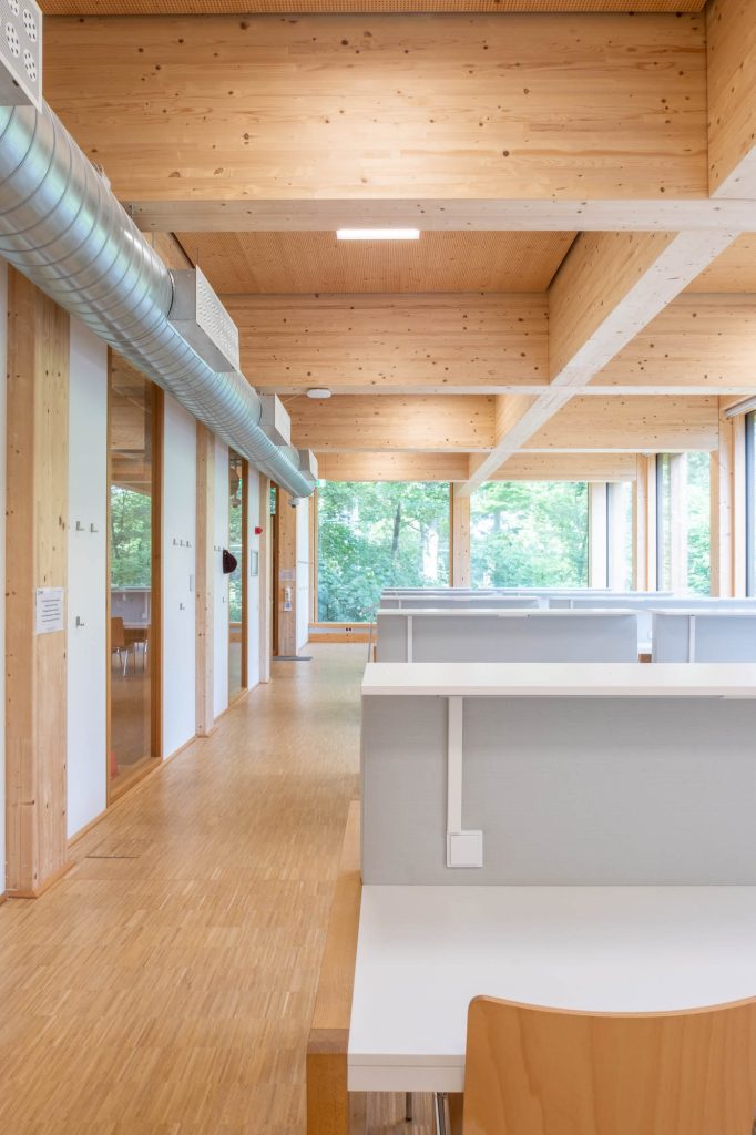A modern, bright study area with wooden beams, light wood flooring, large windows with green trees outside, and individual white cubicles with wooden chairs. Exposed ducts run along the ceiling.