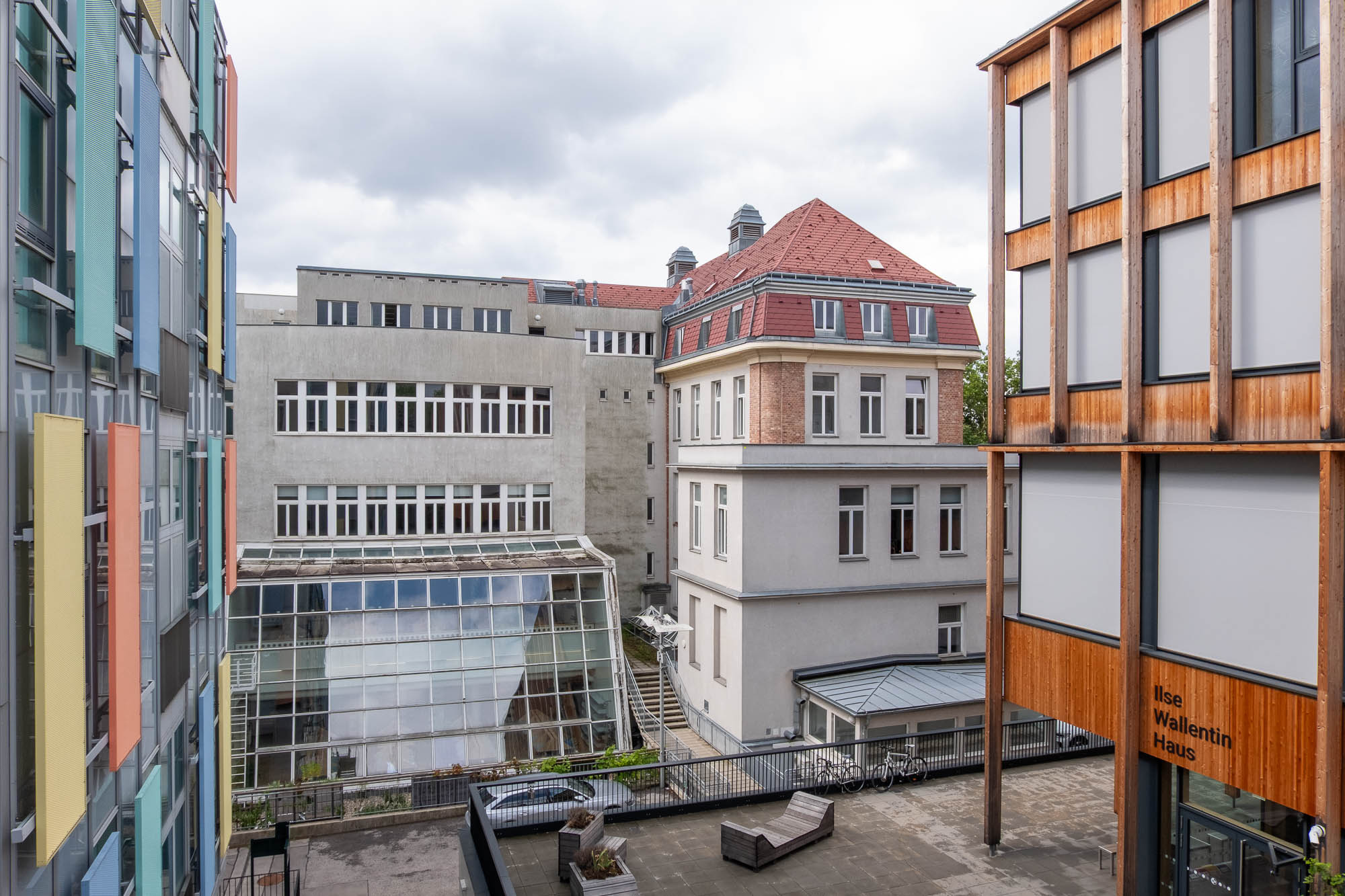 A courtyard bordered by modern and older buildings, featuring colorful panels on the left, glass windows in the center, and a wooden-paneled structure labelled Ilse Wallentin Haus on the right under an overcast sky.
