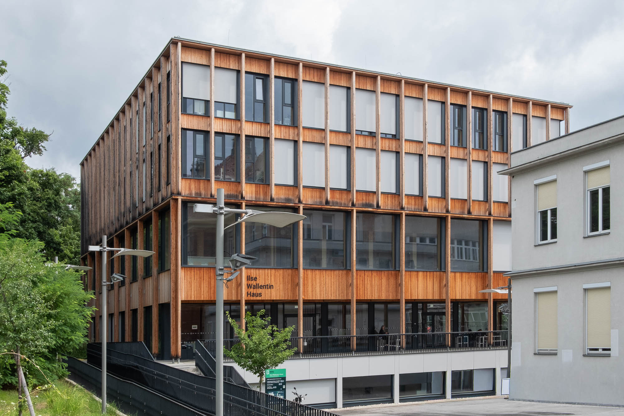 A modern, three-story building with a wooden exterior and large windows. The words "Ilse Wallentin Haus" are displayed on the facade. The sky is overcast and a few trees and street lamps are visible.
