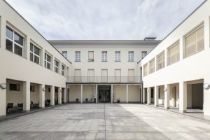 A rectangular courtyard with paved stone flooring is surrounded by a three-story cream-colored building with multiple windows and covered walkways.