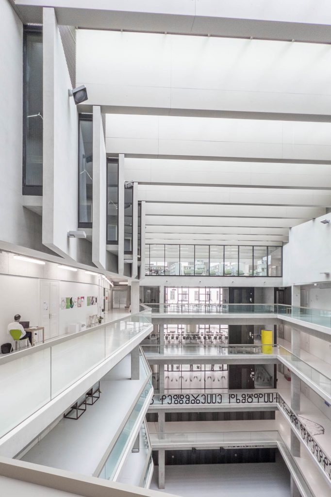 A modern, multi-story atrium in a building with white walls, glass railings, and open walkways. The area is well-lit with natural light from large windows and skylights. A few people are visible on the walkways.