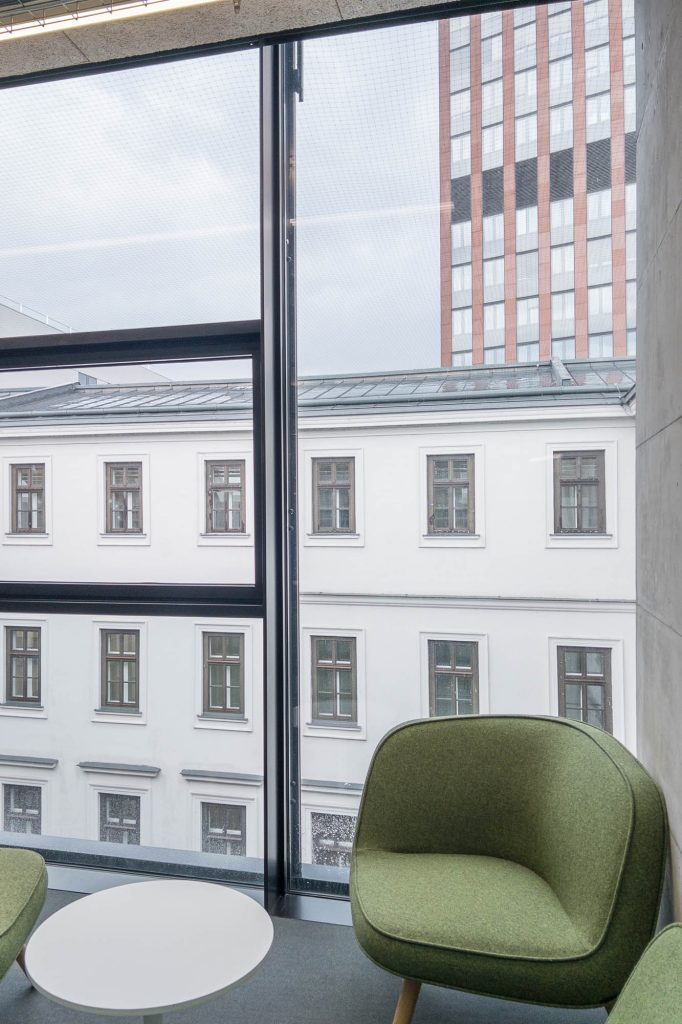 Two green chairs and a small round white table are placed by a large window overlooking white and red buildings.