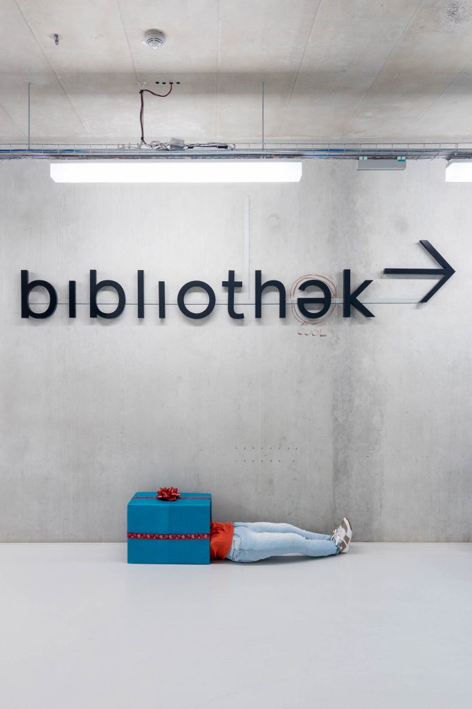 A mannequin lies on the floor with its upper body hidden behind a large blue gift box with a red ribbon, against a concrete wall with the word bibliothek and an arrow sign.