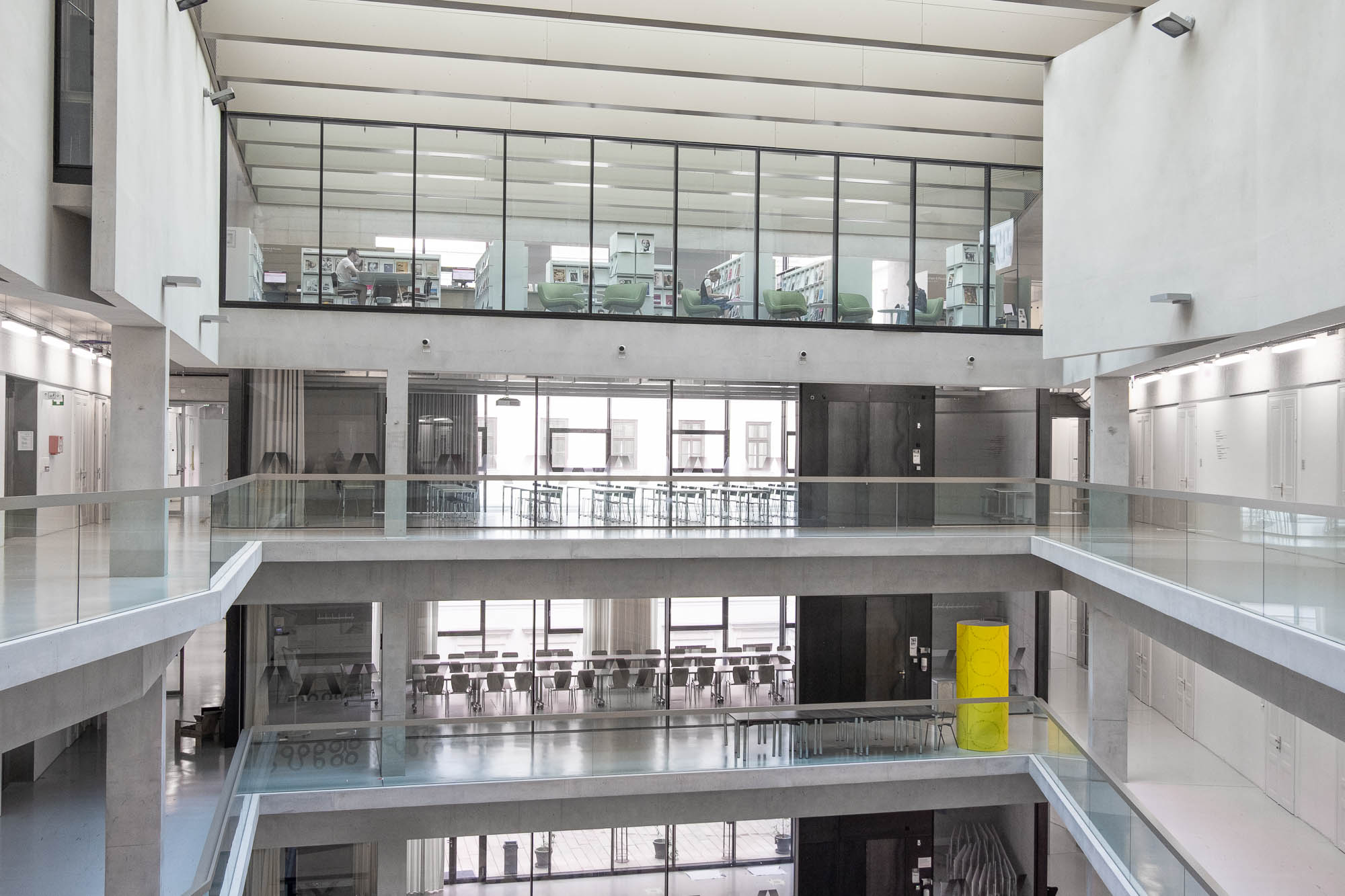 A modern multi-story building interior with large glass windows, open walkways, and visible tables and chairs. The space appears bright with natural light and has a minimalist design featuring concrete and glass elements.