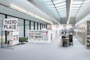 A modern, brightly lit library or office space with shelves of magazines, a green chair, workstations with computers, and a sign reading “THIRD PLACE” near large glass windows.