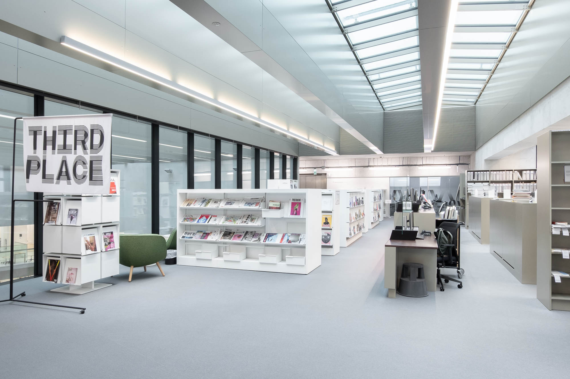 A modern, brightly lit library or office space with shelves of magazines, a green chair, workstations with computers, and a sign reading “THIRD PLACE” near large glass windows.