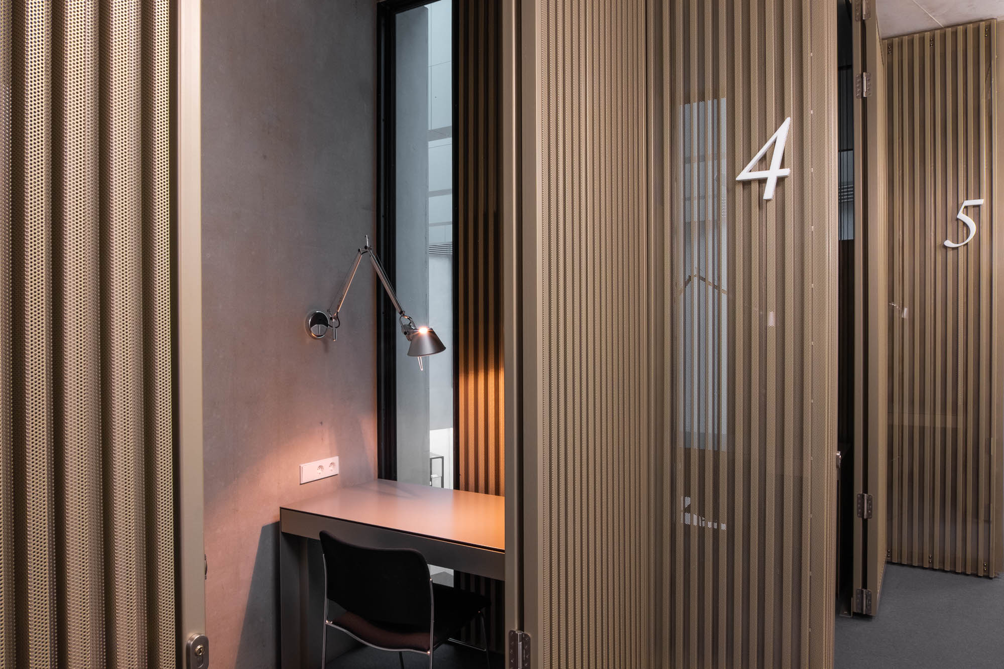 A small study booth with a black chair and desk lamp. The walls are a copper-coloured lattice of perforated steel and the numbers 4 and 5 are affixed next to the doors.