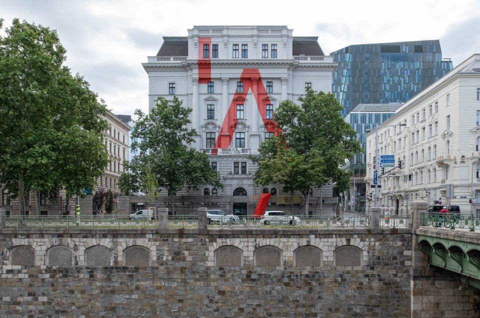 A white Neoclassical building stands between trees on a riverbank. The facade is painted white with a large stylized red letter A.