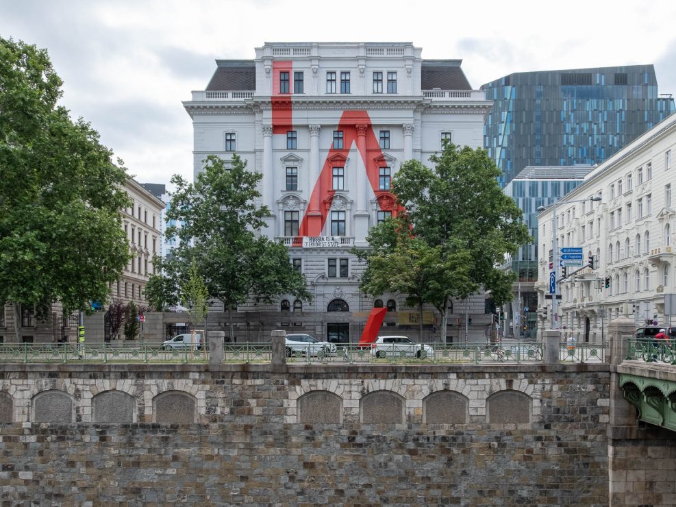 A white Neoclassical building stands between trees on a riverbank. The facade is painted white with a large stylized red letter A.