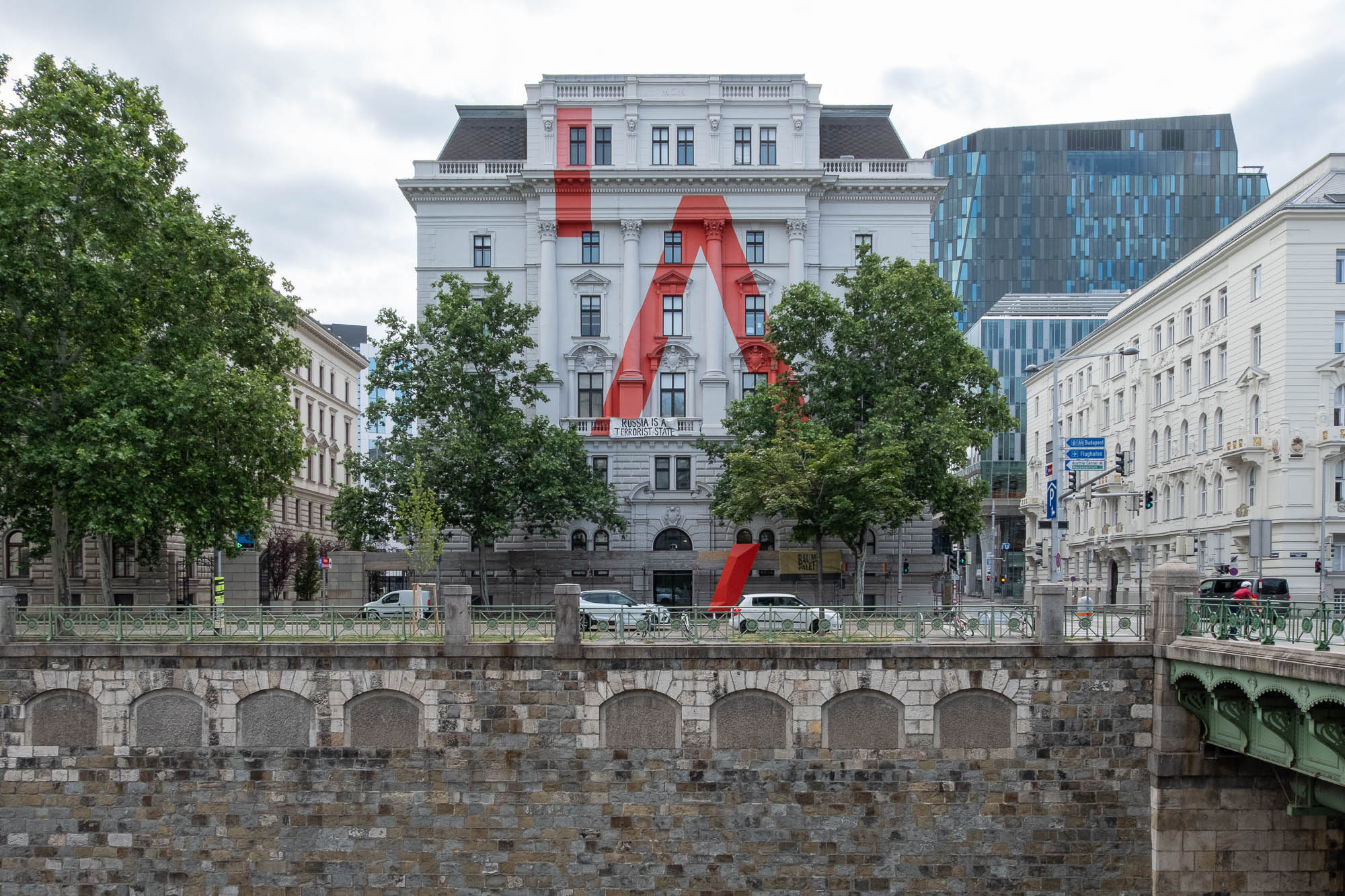 A white Neoclassical building stands between trees on a riverbank. The facade is painted white with a large stylized red letter A.