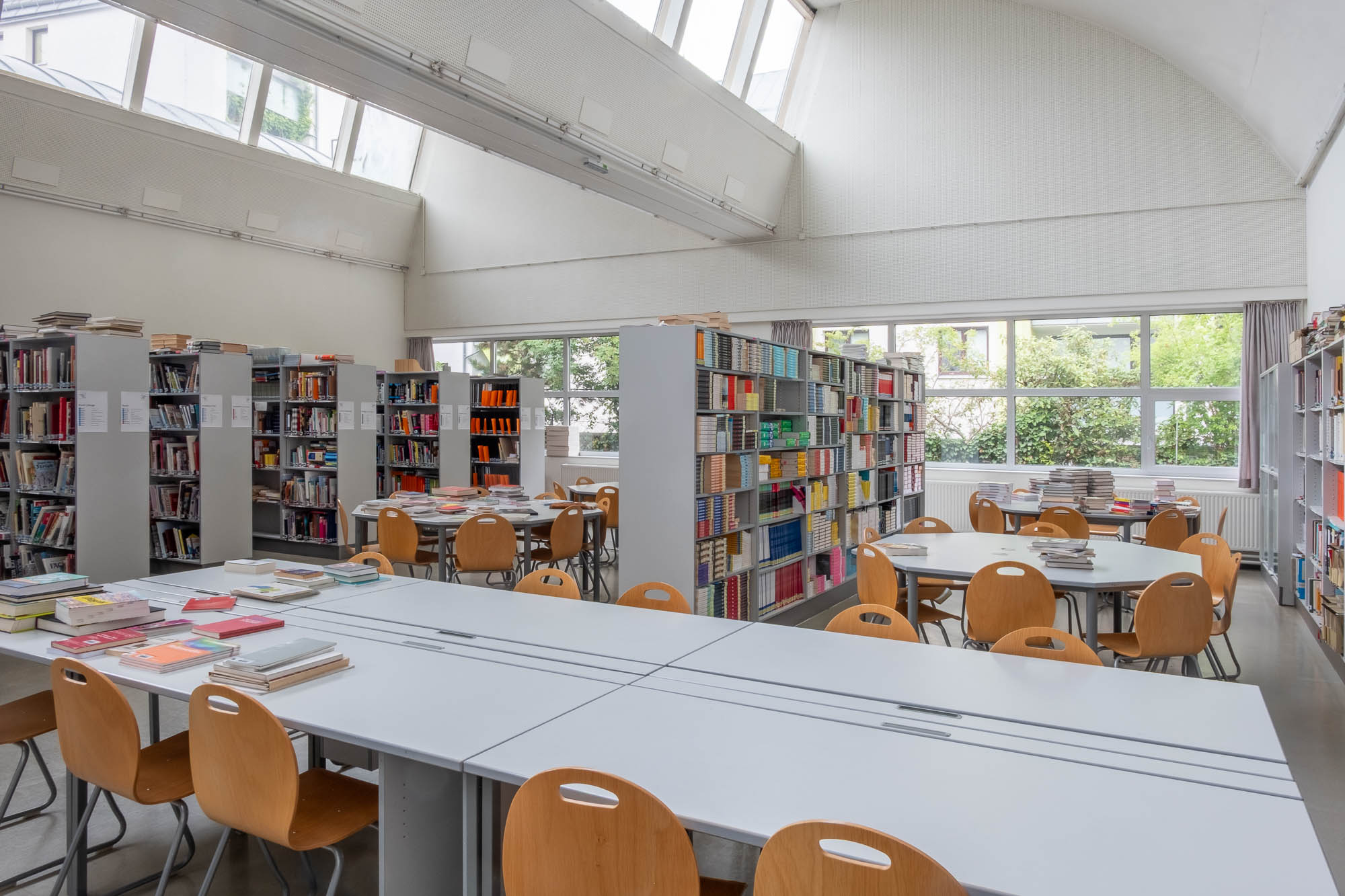 A brightly lit library room with large windows, several bookshelves filled with books, wooden chairs, and white tables arranged for study or reading. Some books and papers are placed on the tables and shelves.
