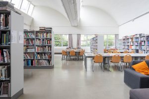 A library interior with bookshelves filled with books, tables and wooden chairs arranged for reading or studying, large windows letting in natural light, and a gray sofa with orange pillows.