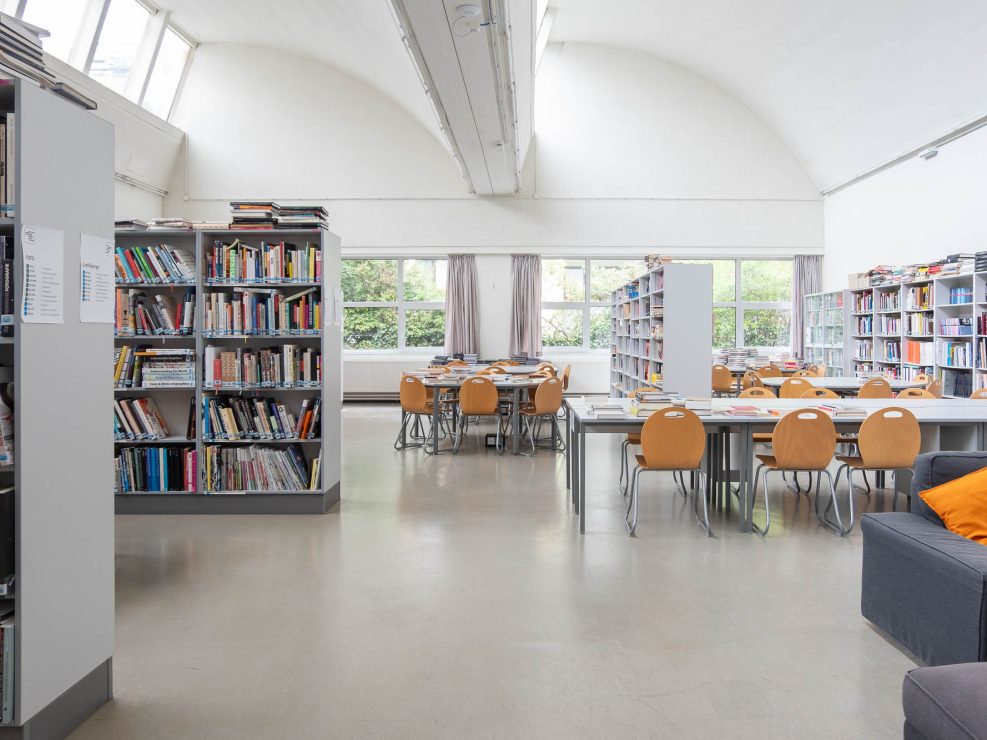 A library interior with bookshelves filled with books, tables and wooden chairs arranged for reading or studying, large windows letting in natural light, and a gray sofa with orange pillows.