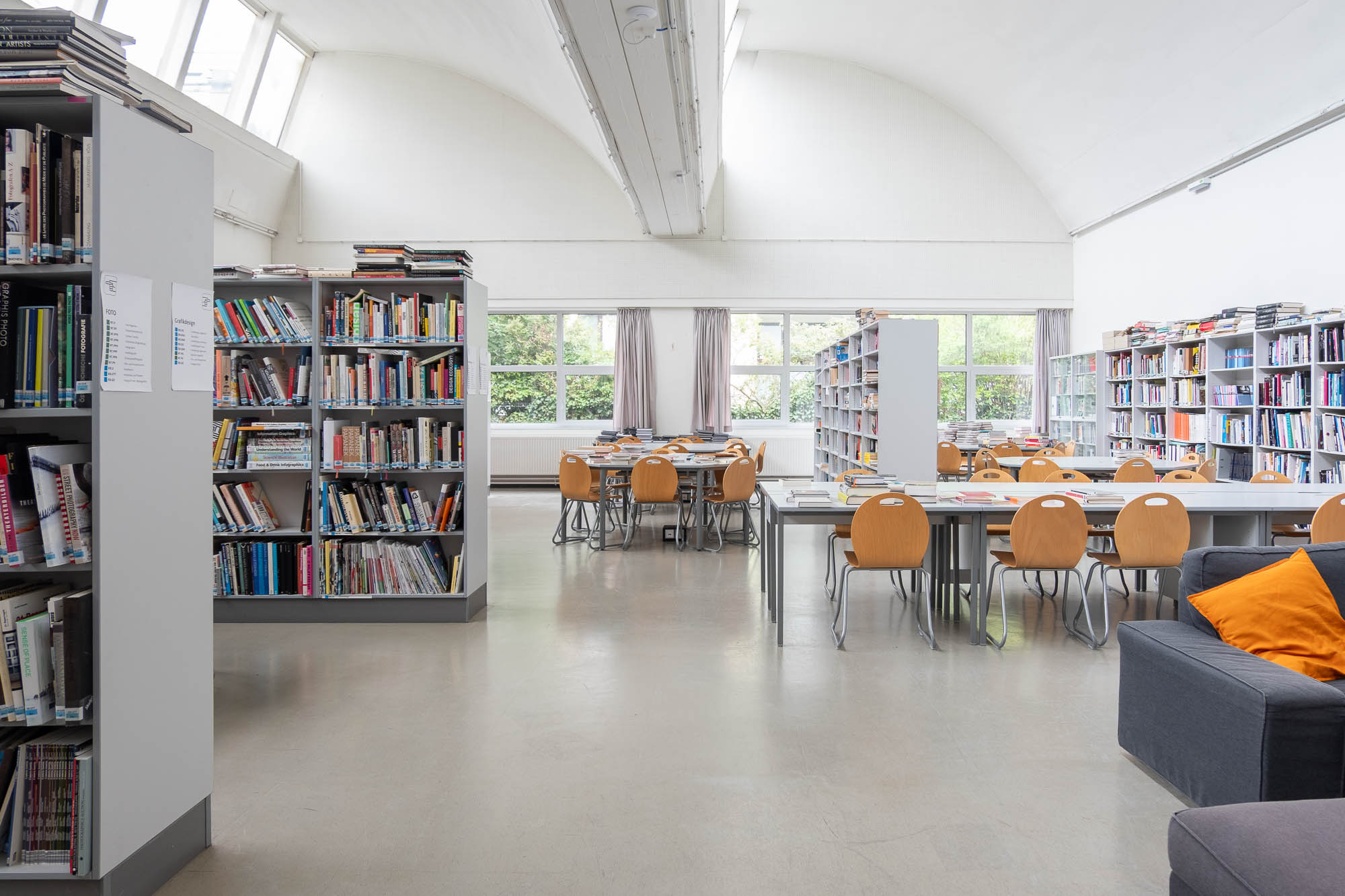 A library interior with bookshelves filled with books, tables and wooden chairs arranged for reading or studying, large windows letting in natural light, and a gray sofa with orange pillows.