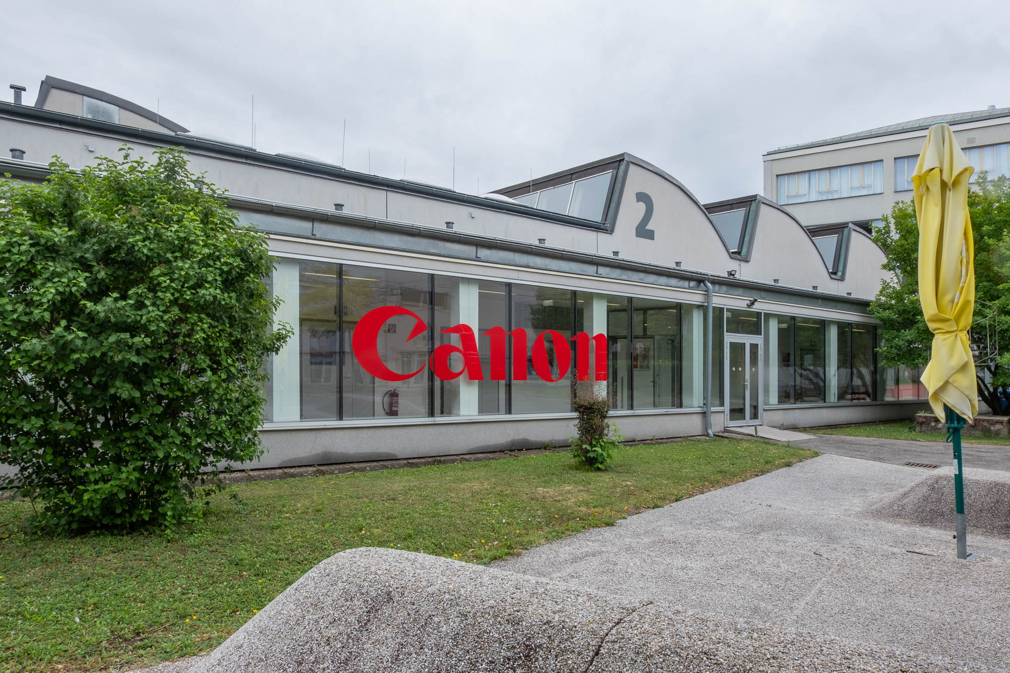 A building with large windows displays a prominent red Canon logo. The building is marked with the number 2, surrounded by grass, bushes, and an unopened yellow umbrella on the right side.
