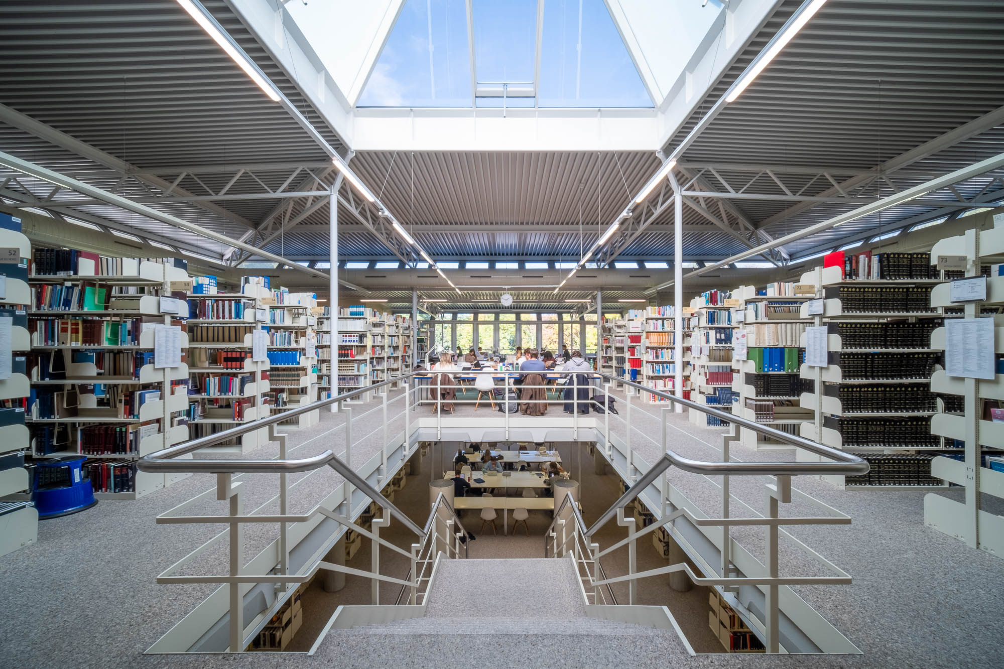 A modern library interior with bookshelves on both sides, a central staircase leading down to a study area, and large windows and a skylight allowing natural light to fill the space. People are seated at tables working.