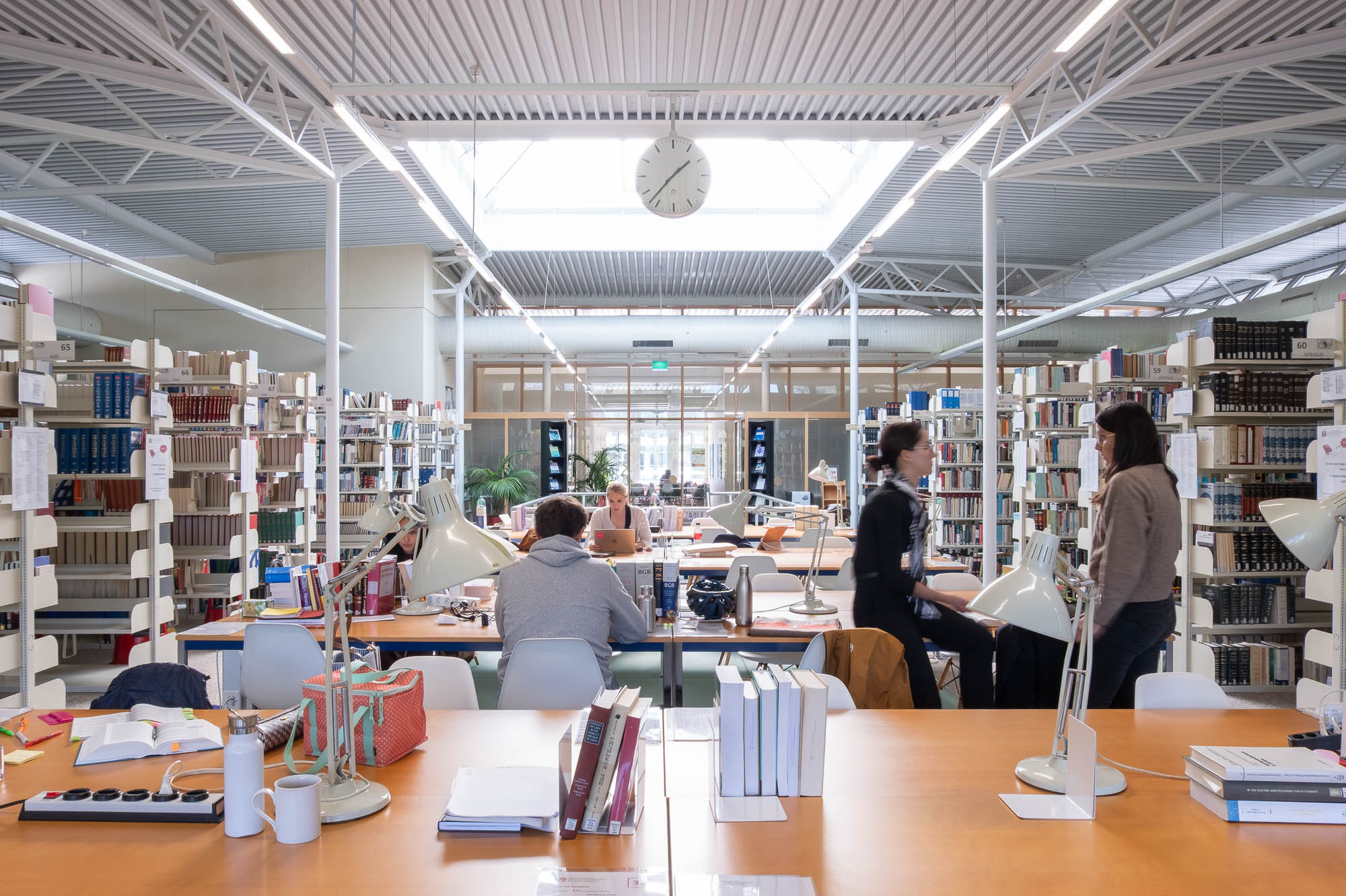 A modern library interior with high ceilings, bookshelves, large windows, and people reading and working at tables. Multiple lamps and books are on the tables, and a clock hangs from the ceiling at the center of the room in front of a skylight.