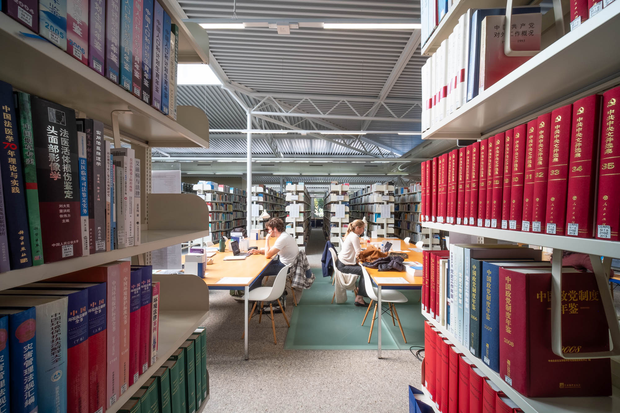 People sit and study at tables in a library, surrounded by shelves filled with books, some with Chinese text on the spines. The library has a modern, well-lit interior with a high ceiling.
