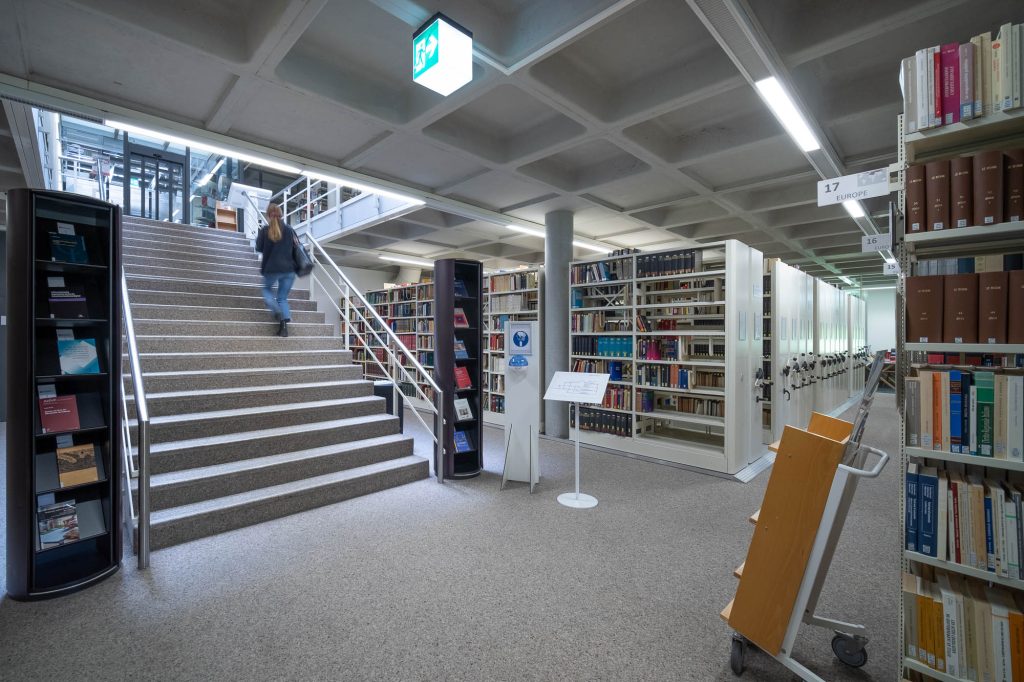 A person ascends a staircase in a modern library filled with bookshelves, book carts, and signage. There's a book cart in the front.