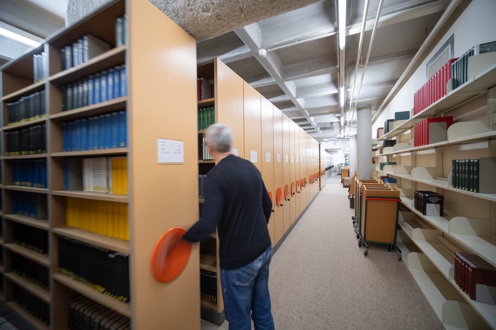A person turns the handle of a movable shelving unit in a library, with rows of shelves filled with books on both sides and carts for transporting books along the aisle.