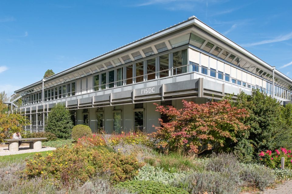 A two-story modern building with large windows and a gray roof, surrounded by landscaped gardens and bushes. The text "l'ISDC" is written on the building.