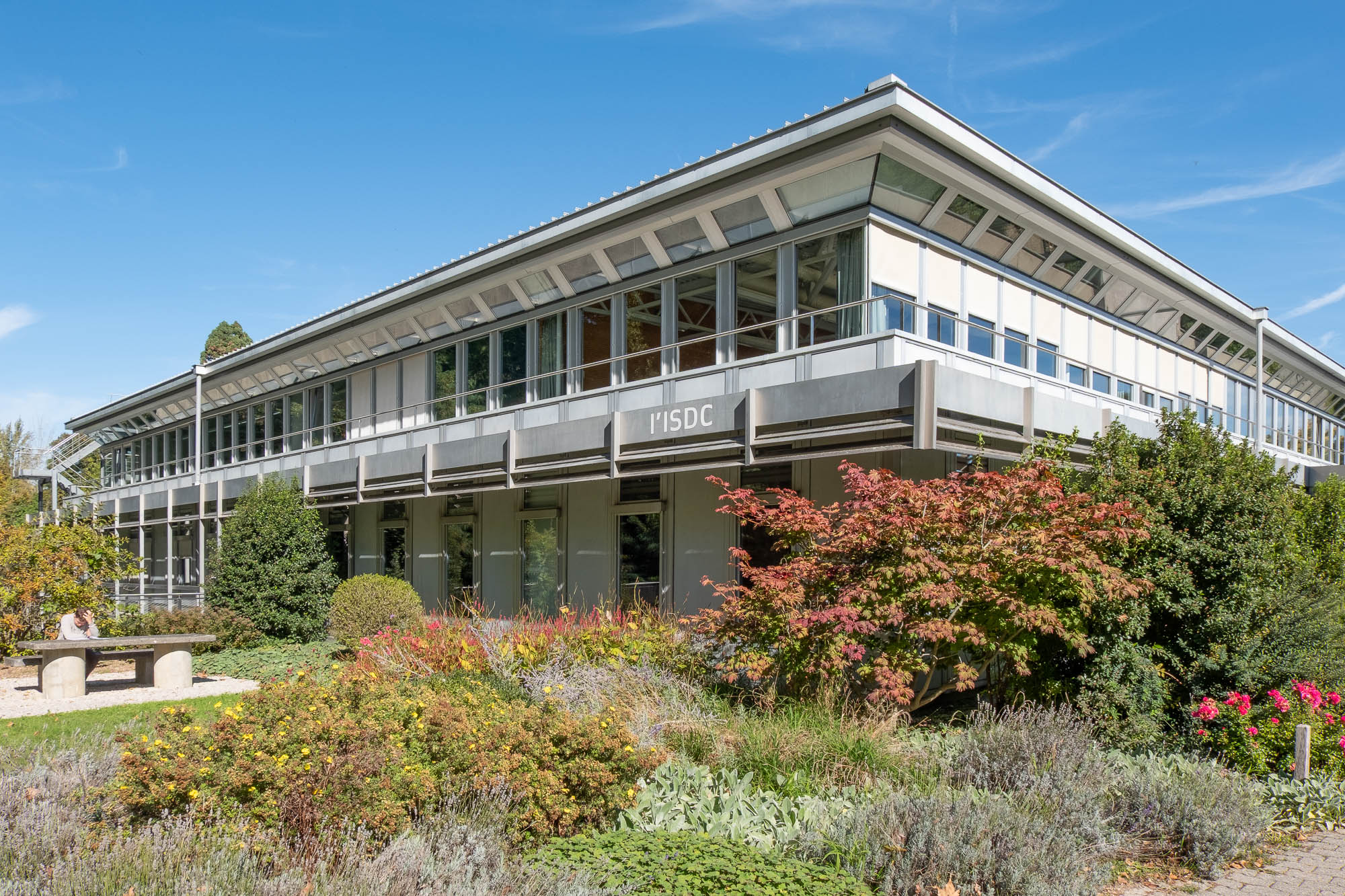 A two-story modern building with large windows and a gray roof, surrounded by landscaped gardens and bushes. The text "l'ISDC" is written on the building.