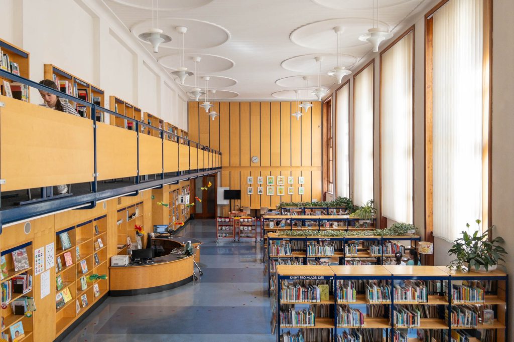 A spacious, modern library with tall windows, wooden bookshelves filled with books, a front desk, and a second-floor balcony. Potted plants are placed on shelves, and a person is reading on the upper level.