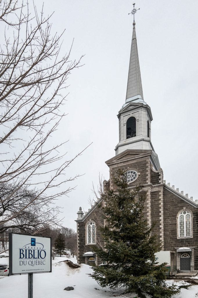 Exterior of a stone church in winter. A sign reading "Réseau biblio du Québec" stands in front.