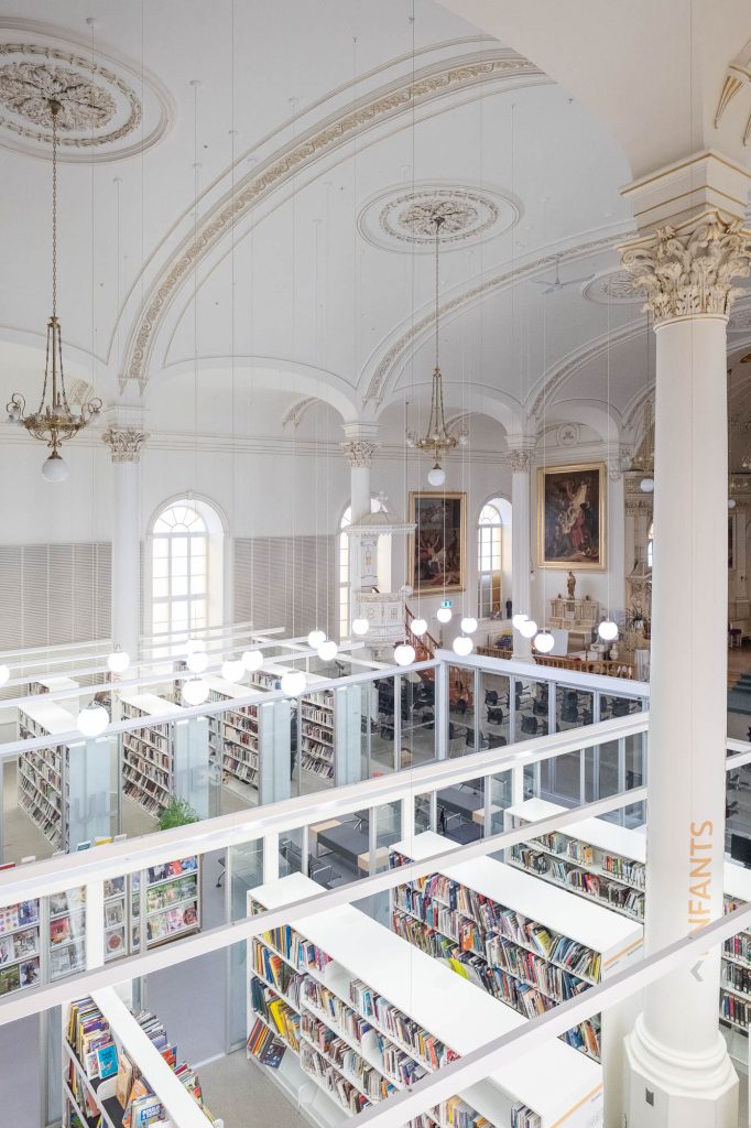 Interior of a church converted to a library. A glass enclosure containing the library is in the forefront, only occupying part of the church, which can be seen at the back.