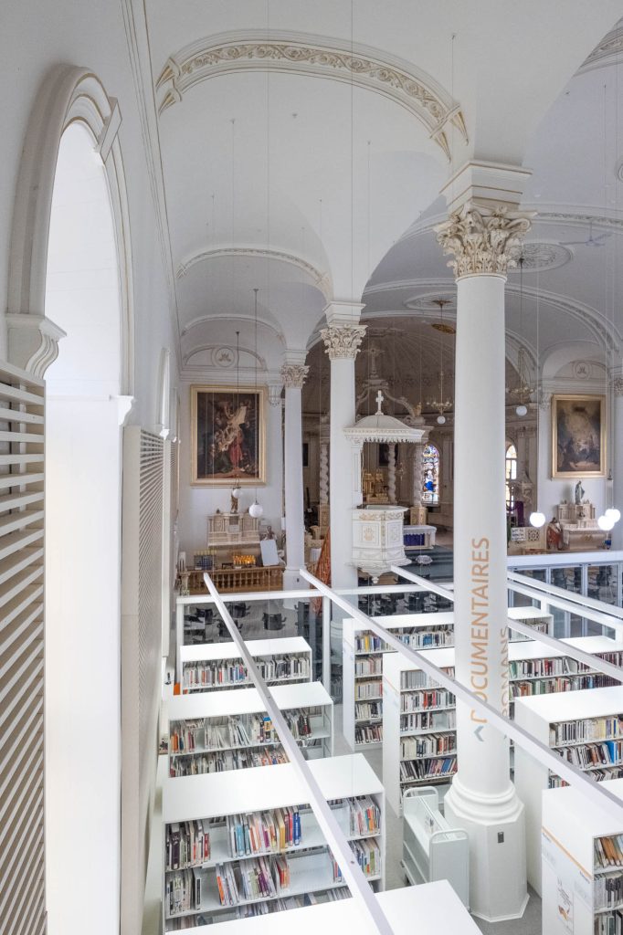 Interior of a church converted to a library. A glass enclosure containing the library is in the forefront, only occupying part of the church, which can be seen at the back.
