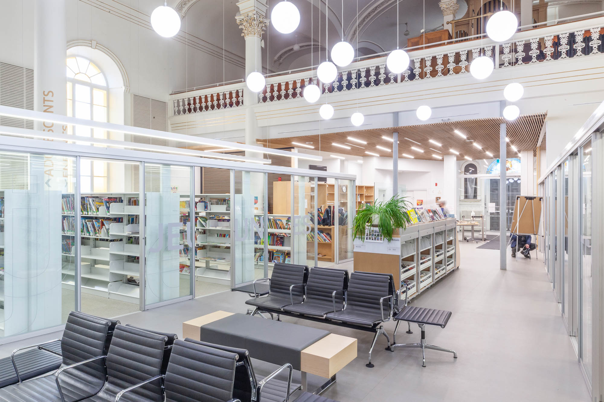 Interior of a church converted to a library. The lower floor is occupied by a contemporary library with modern furniture, glass walls and globe lights. The original church balcony with an organ can be seen above.