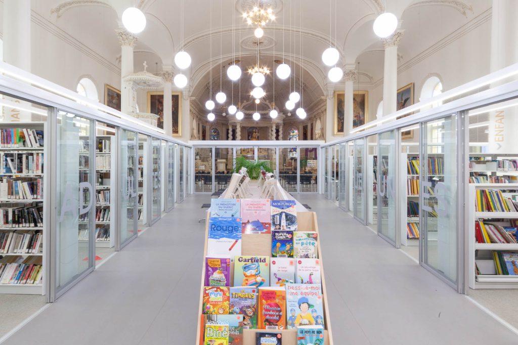 Interior of a church converted to a library. A glass enclosure containing the library is in the forefront, only occupying part of the church, which can be seen at the back.