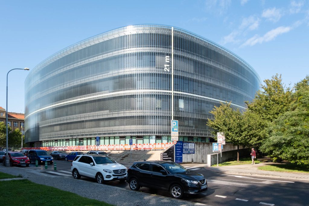 A modern, oval-shaped glass library building on a quiet street lined with parked cars. White lines indicating measurements are painted on the building, the vertical line indicates 21m. A person walks through a wooded park to the right.