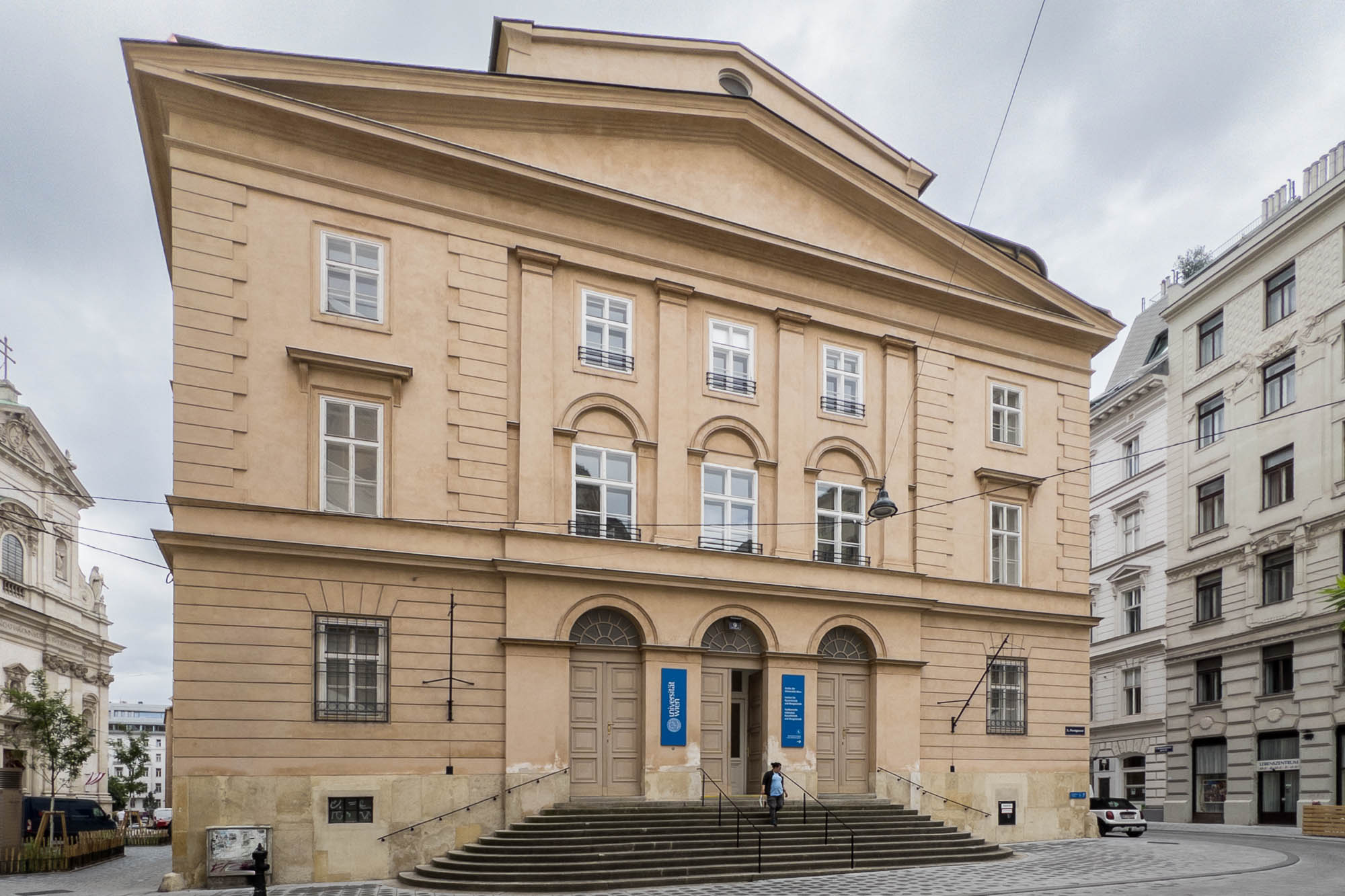 A large beige neoclassical building with three arched doorways, tall windows, and stairs leading to the entrance. Two blue vertical banners hang by the doors with the logo of the University of Vienna. A person stands on the steps. Other buildings are nearby.