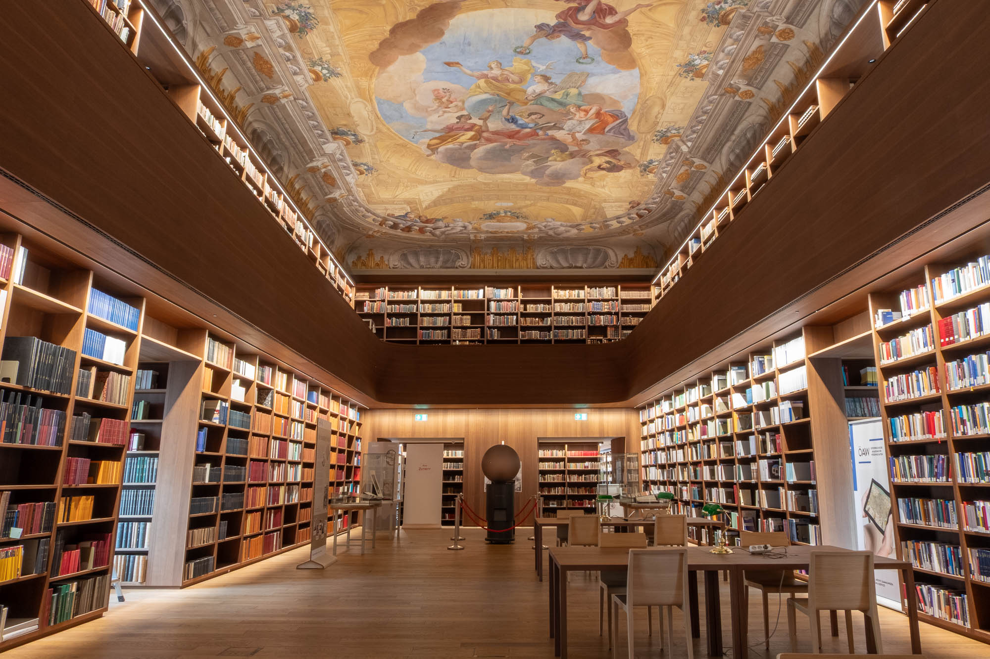 A spacious library with wooden floors, rows of bookshelves filled with books, study tables with chairs, a ladder by the shelves, and a ceiling decorated with a baroque fresco.