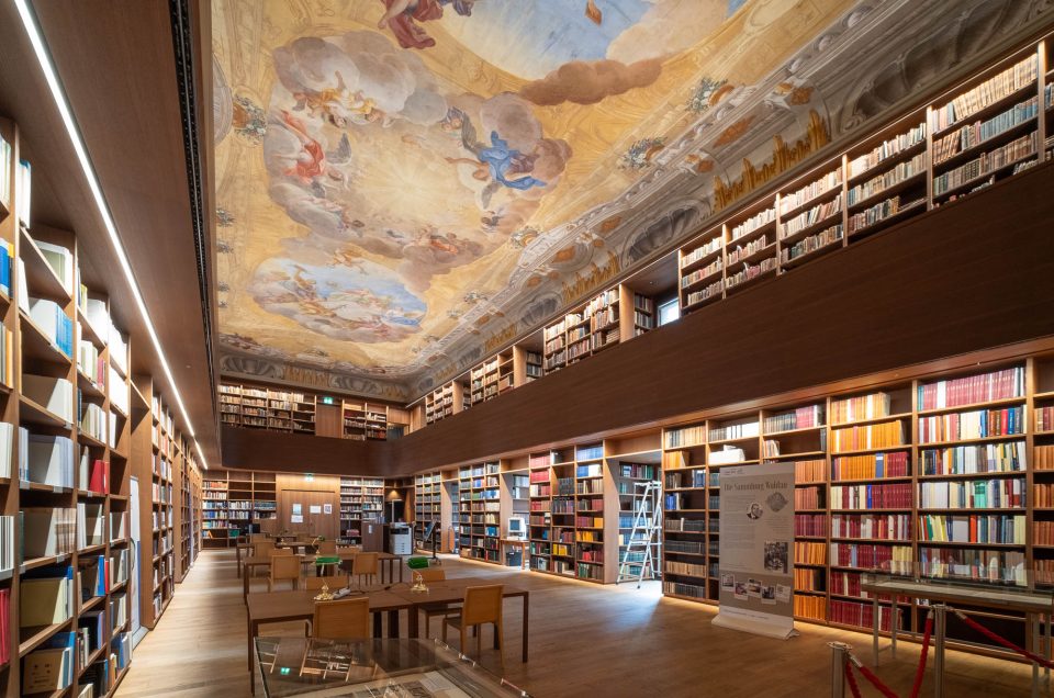 A spacious library with wooden floors, rows of bookshelves filled with books, study tables with chairs, a ladder by the shelves, and a ceiling decorated with a baroque fresco.