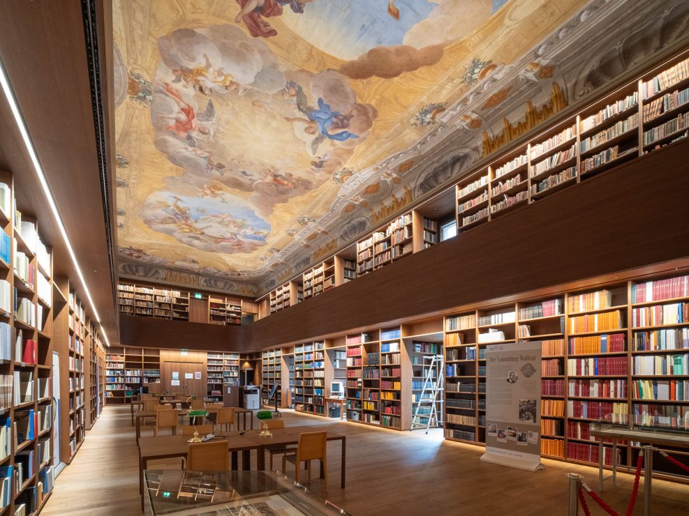A spacious library with wooden floors, rows of bookshelves filled with books, study tables with chairs, a ladder by the shelves, and a ceiling decorated with a baroque fresco.